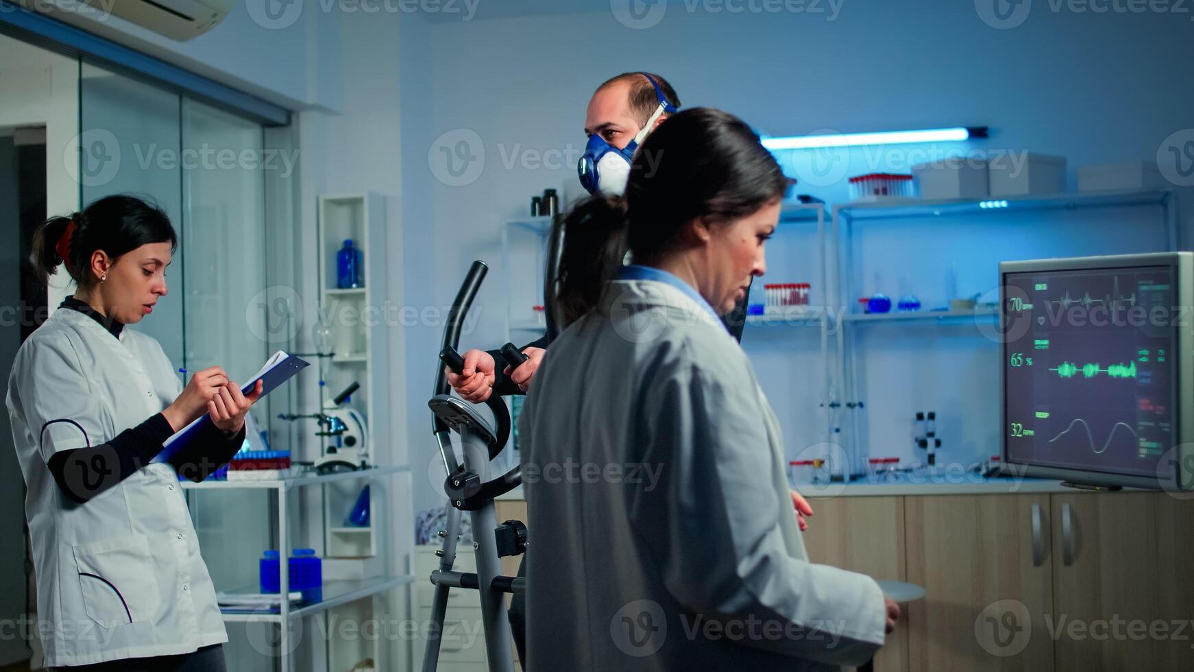 Doctor sport scientist looking at monitor with EKG reading while man with mask running on cross trainer monitoring his endurance. Team of researchers monitoring his endurance and heart rate photo