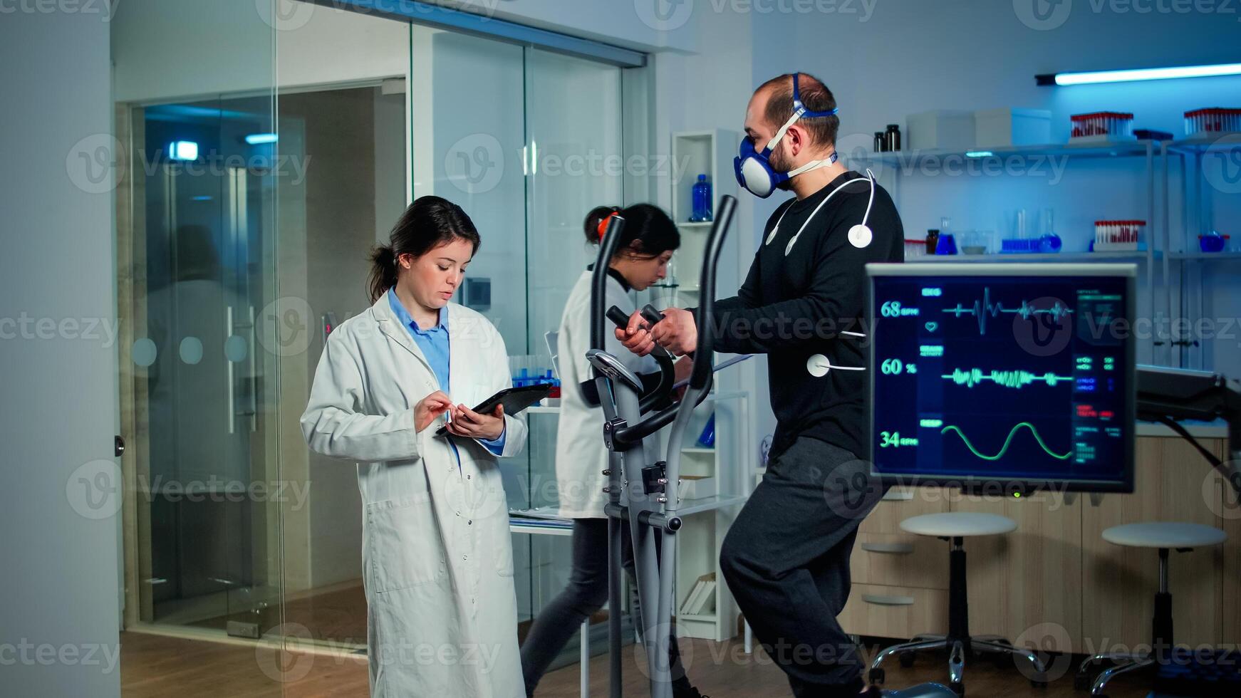 Sportsman with mask running on cross trainer in sports science laboratory measuring VO2 max, heart rate, psychological resistance and muscular endurance. Monitor showing EKG reading of sportsman photo