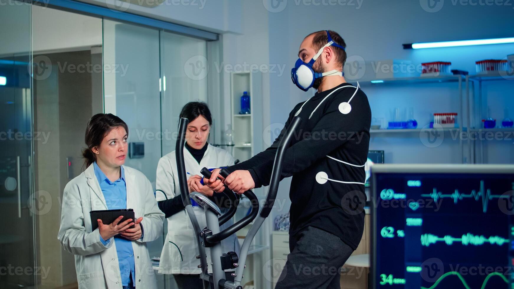 Team of medical researchers monitoring performance athlete with mask and electrodes attached on him measuring endurance, heart rate, psychological resistance, training muscle mass in science sport lab photo