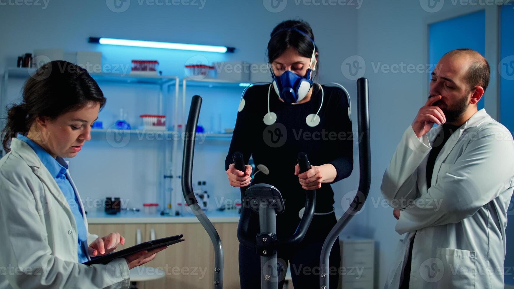 Woman athlete with mask doing fitness exercises in science sport lab with electrodes attached while scientist holding tablet supervising whole process. Physician using notepad controling EKG data photo