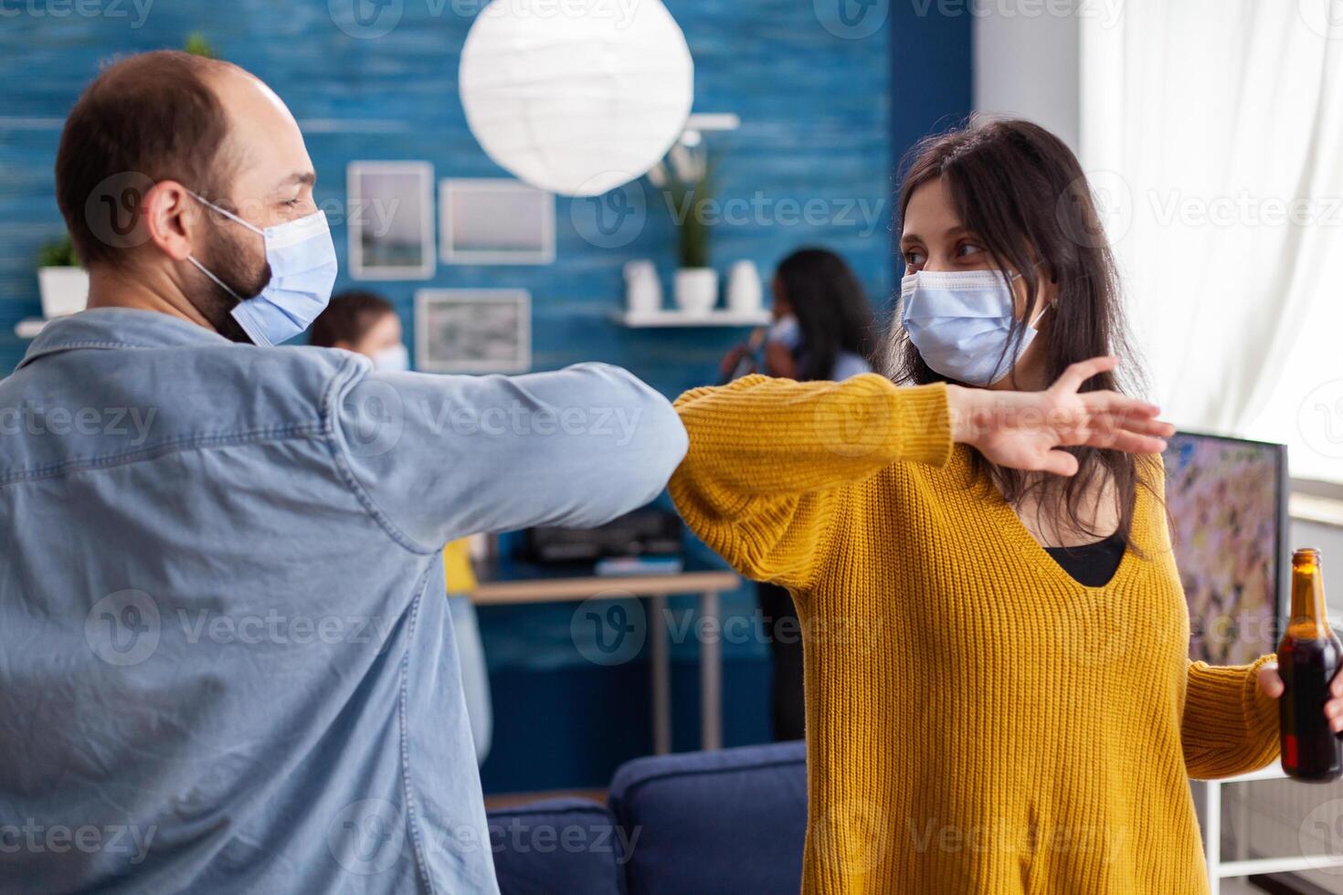 Happy cheerful friends touching elbow keeping social distancing greeting eachother wearing face mask preventing spread of coronavirus holding beer bottles in apartment living room. Conceptual image. photo