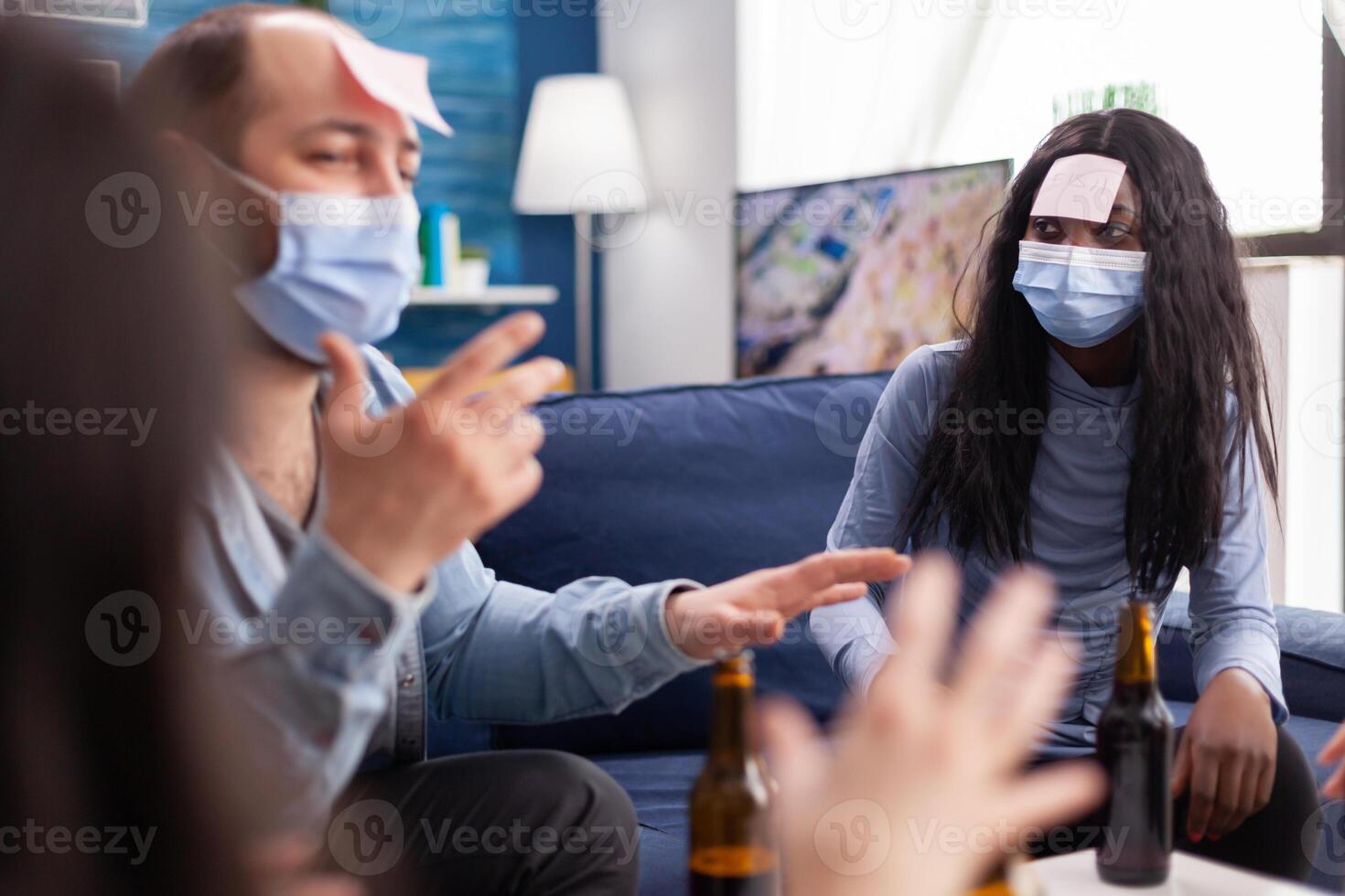 African woman with sticky note on forehead playing name game with cheerful happy friends wearing face mask to prevent infection with covid19 due to social pandemic. Beer bottle, photo