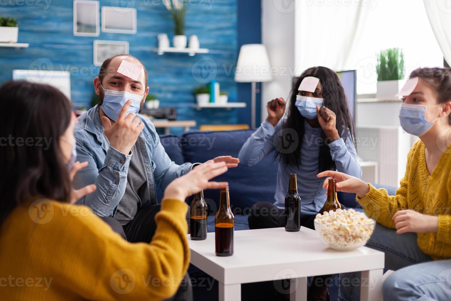 Cheerful friends with sticky notes on foreheads playing name game with beer popcron keeping social distacing with face mask to prevent spread of covid19 in home living room. Conceputal image. photo