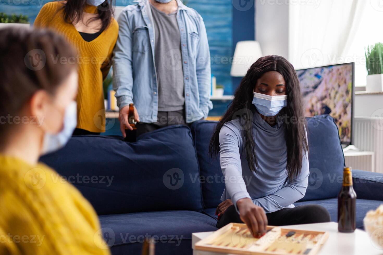 African woman competing with friends playing backgammon wearing face mask keeping social distancing during social pandemic with covid19 drinking beer in home living room. Board games, recreation, players. Conceptual image. photo