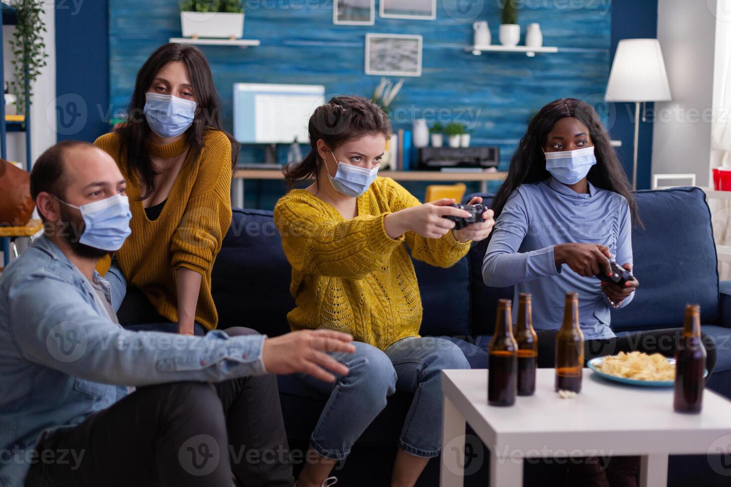Concentrated multiethnic friends trying to win at games during social pandemic wearing face mask keeping distancing not to spread corona sitting on couch in living room. photo