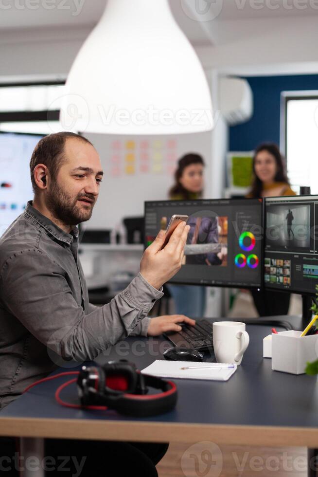 editor discussing online with customer designer holding smartphone editing footage sitting in multimedia company. Man creator artist processing movie montage using post production software photo
