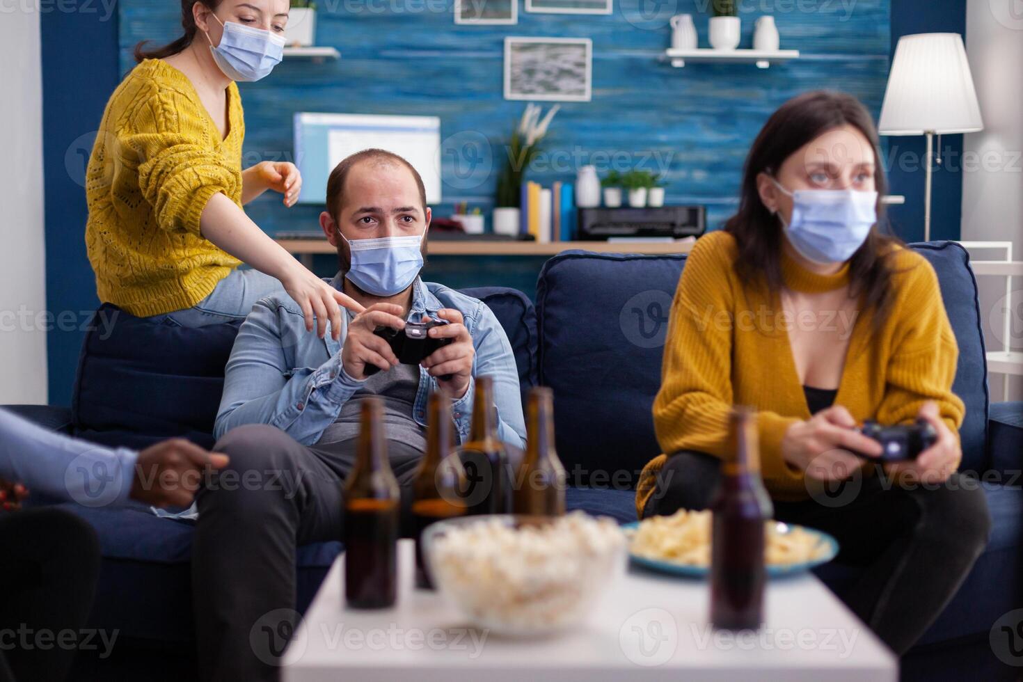 Diverse friends trying to win playing games using joystick havin fun wearing face mask to prevent spreading of coronavirus in time of global outbreak. Gaming competition, beer and popcorn. photo