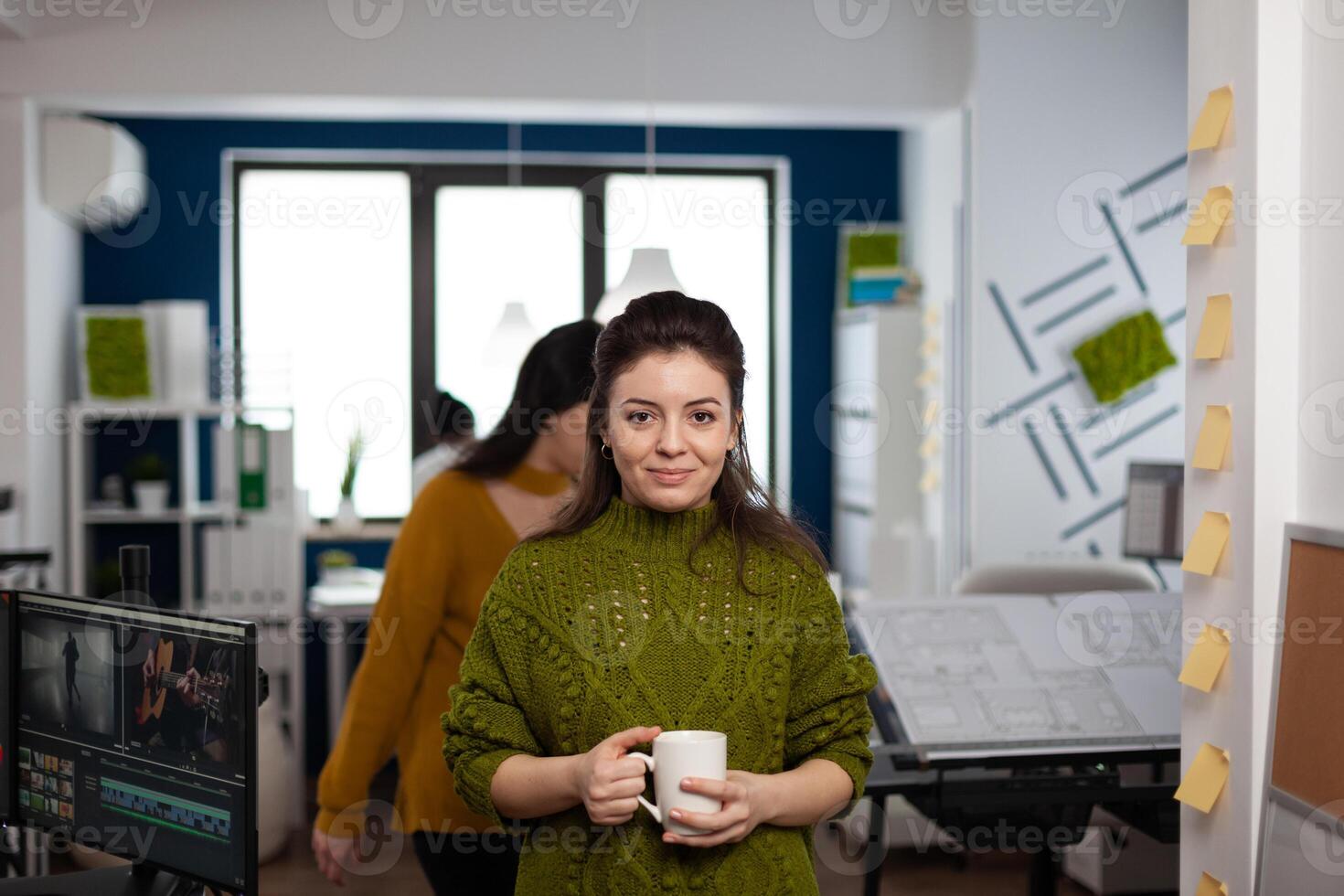 Portrait of designer artist smiling at camera holding cup of coffee standing in start up multimedia agency office. Woman working in creativity studio production editing design with diverse team. photo
