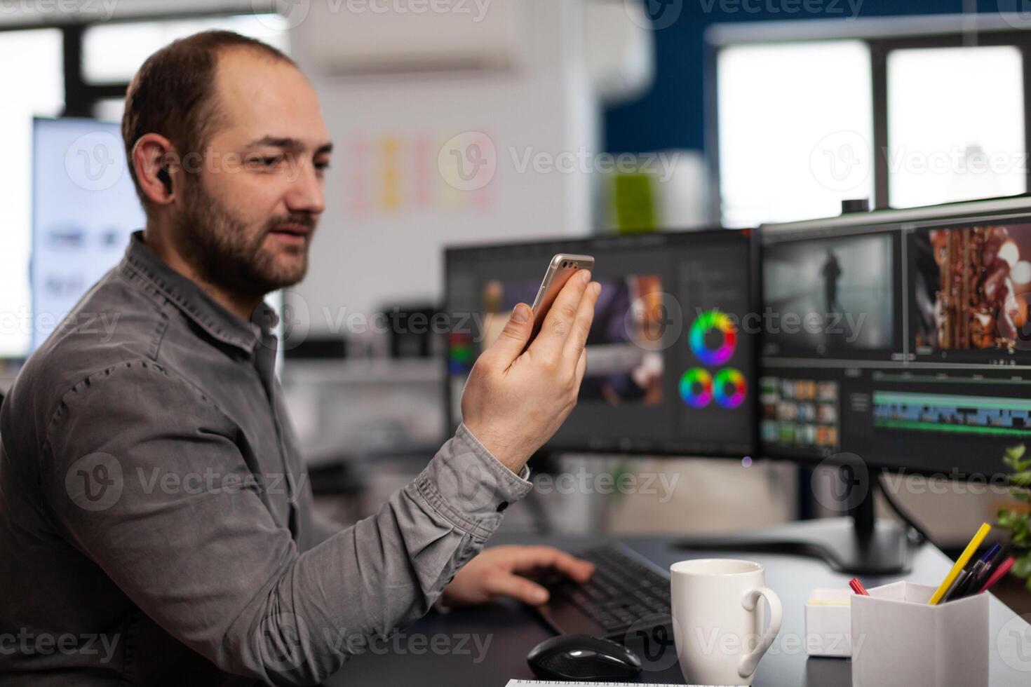 Multimedia creator artist talking on videocall with client holding smartphone editing footage sitting in creativity production agency. Videographer using post production software photo