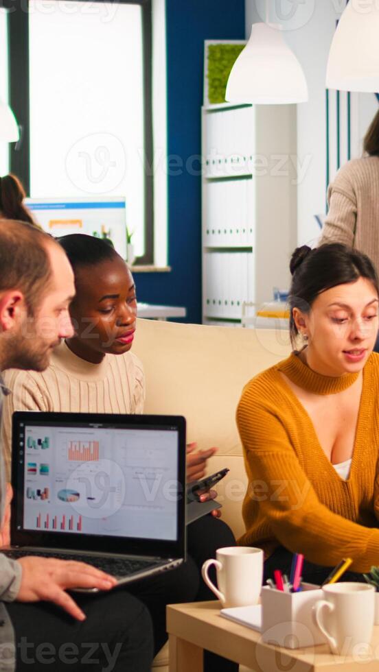 Businesswoman corporate leader talking to diverse team explaining project plan reviewing paperwork sitting on couch. Multiethnic coworkers taking notes on laptop and tablet analysing financial reports photo