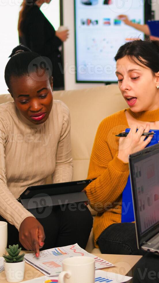 Confident company manager giving working tasks to diverse teammates sitting on couch in start up office. Multiethnic team discussing project ideas at brainstorming meeting using digital devices photo