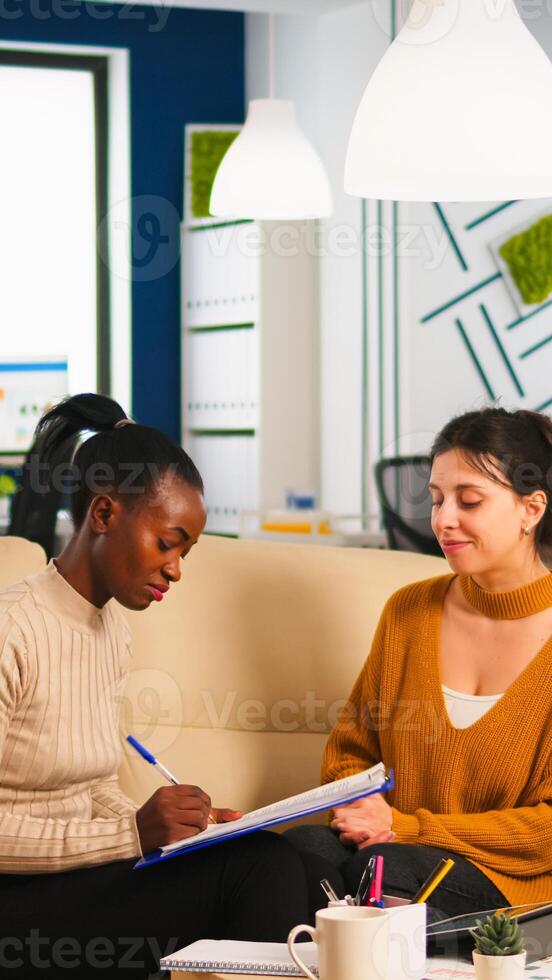 Diverse businesswomen discussing new project for company evolution, black woman checking tasks on clipboard, signing new contract. Multiethnic employees gathered in co-working space photo