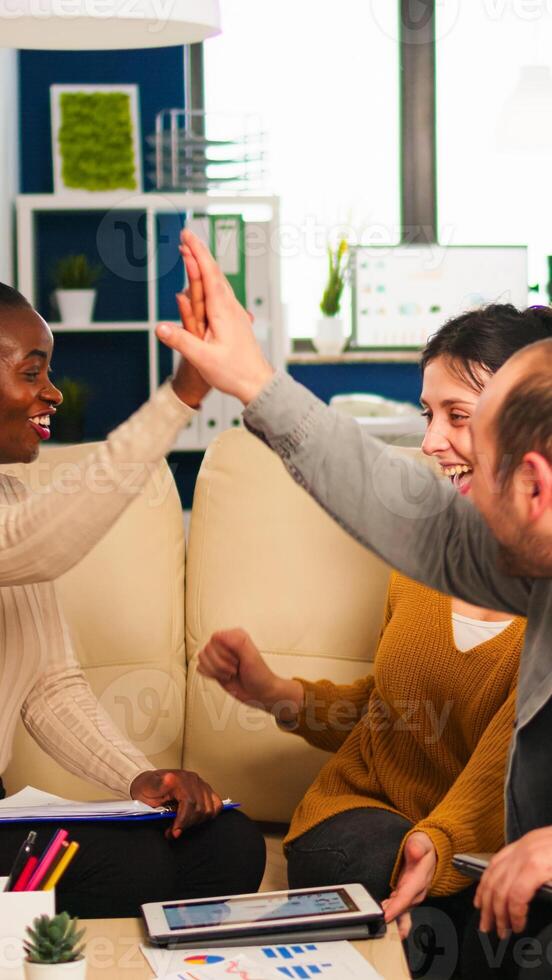 Diverse team of successful young entrepreneurs sharing high-five celebrating success sitting on couch in start up office room. Multiethnic business team with laptop and papers excited about project photo