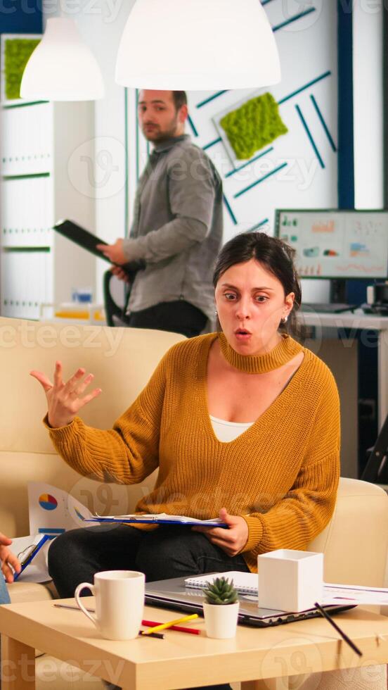 Stressed busineswomen arguing, yealing at each other during work hours sitting on the couch, while diverse colleagues working scared in background. Women throwing papers, conflicts at work concept photo