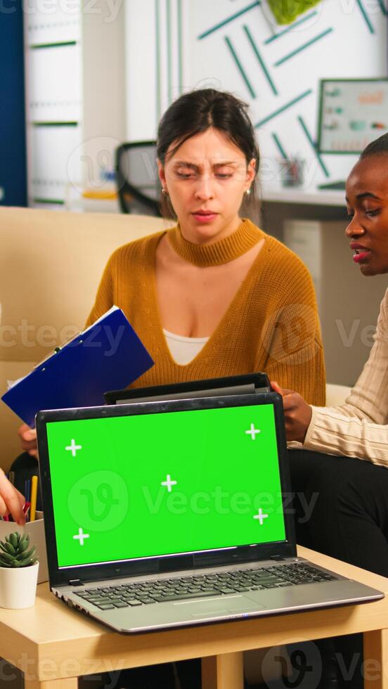Diverse women sitting at table in start up office discussing strategy with green screen laptop in front of camera. Lady explaining to multiethnic team new project, chroma key display. mock up monitor photo