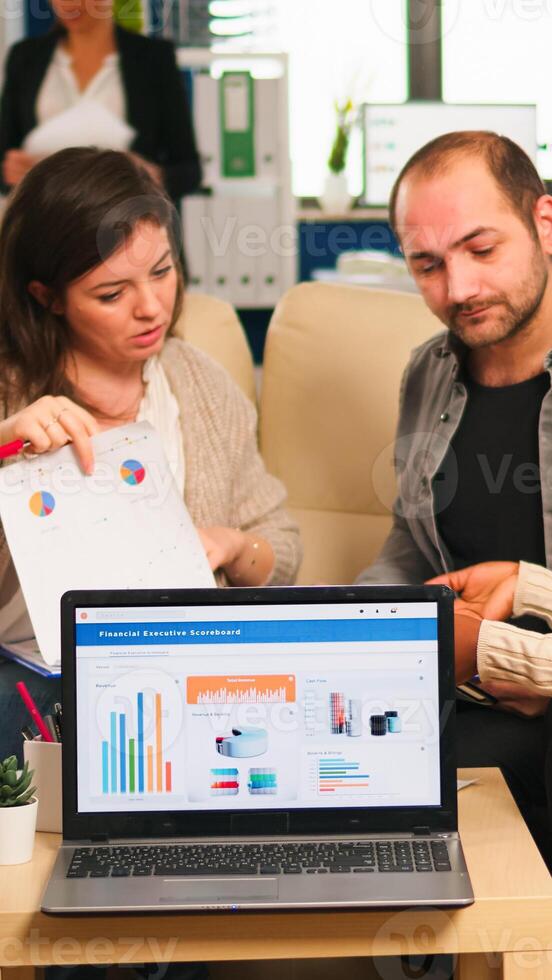 Diverse colleagues employees cooperating in office discussing at workplace, analysing graphs sitting on couch. Business coworkers planning start up business project, brainstorming in corporate office photo