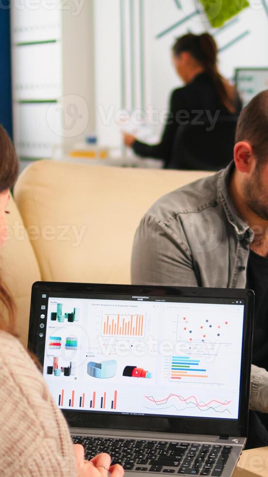 Close up of back view young entrepreneur sitting on couch in office using laptop computer while colleagues working on background. Multiethnic coworkers planning new financial project in modern company photo