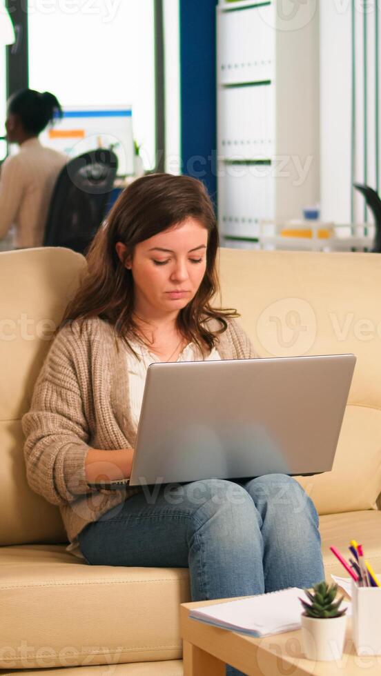 Employee holding laptop sitting in relax zone on confortable couch typing on pc smiling while diverse colleagues working in background. Multiethnic coworkers planning new financial project in company photo
