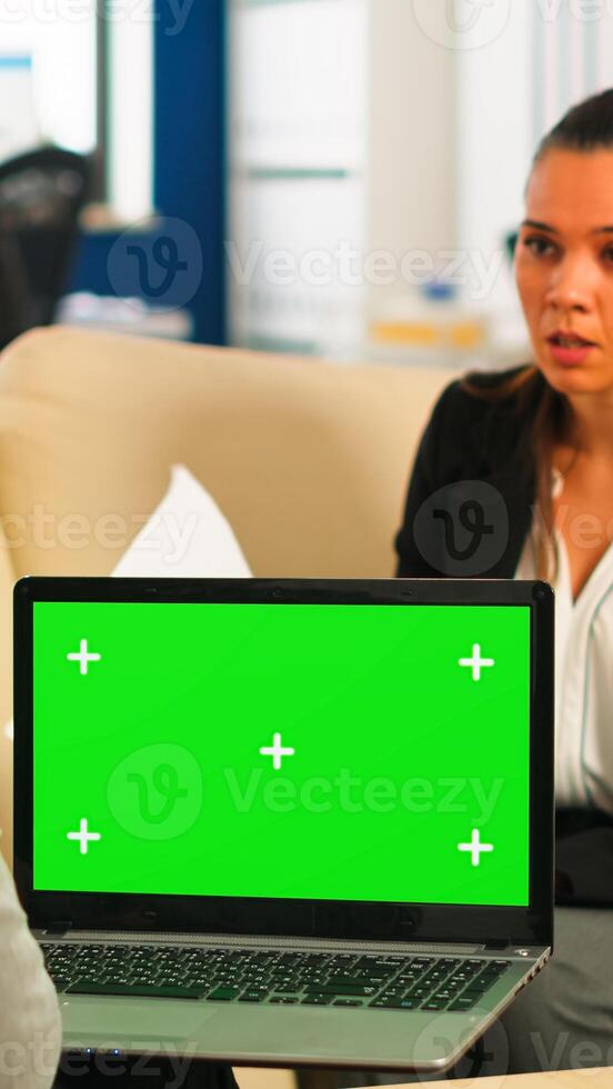 Business team sitting on couch analysing financial statistics, holding laptop with green screen while diverse team working on background. Multiethnic coworkers planning project on chroma key display photo