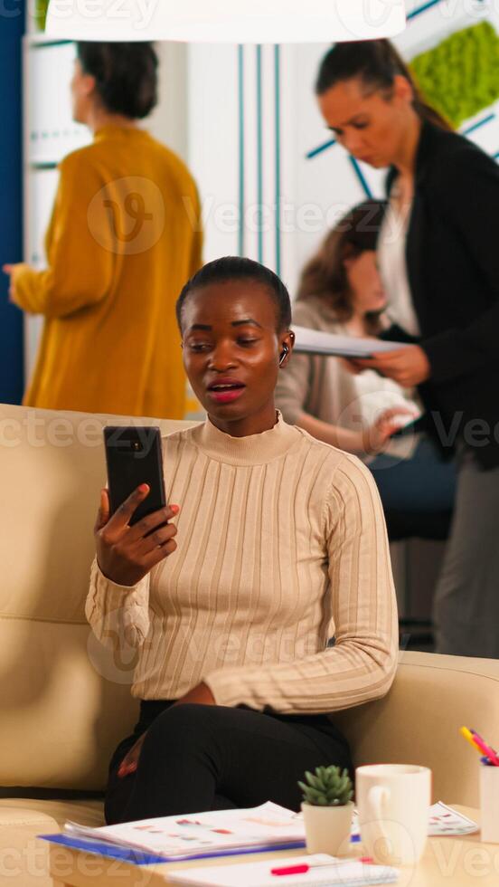 Tired black woman talking with colleague using call holding smartphone with wiraless headset sitting on couch in business office. Diverse team planning new financial project in modern company photo