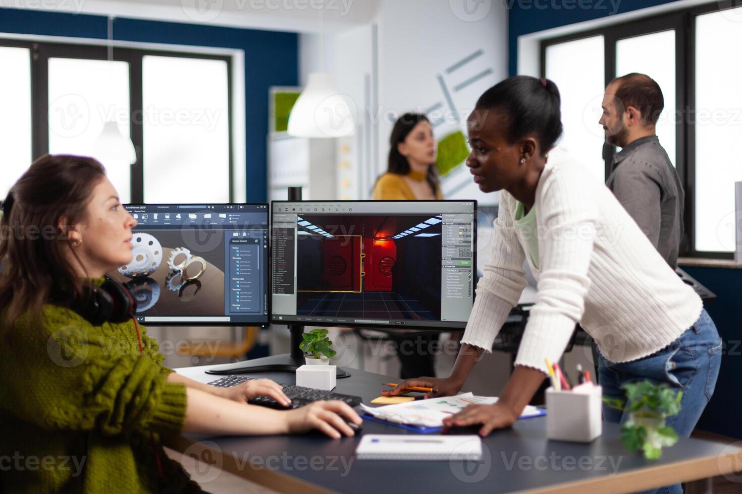 Multiethnic women designer looking at computer with dual displays working together at gaming project in company office. Gamer workers developing new games application on pc with modern technology photo