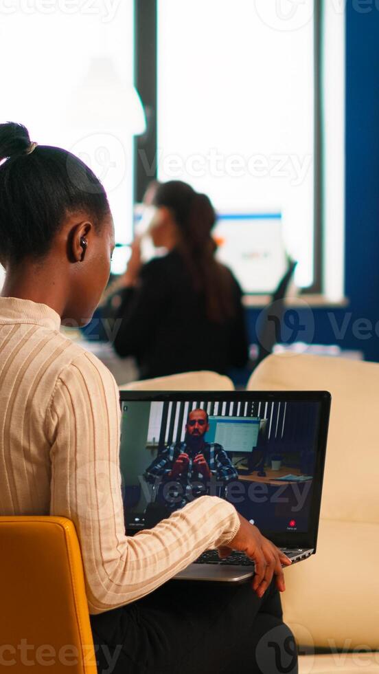 Black woman discussing with remote disabled colleague using call holding laptop sitting on couch in business modern office. Diverse coworkers planning new financial project in modern company photo