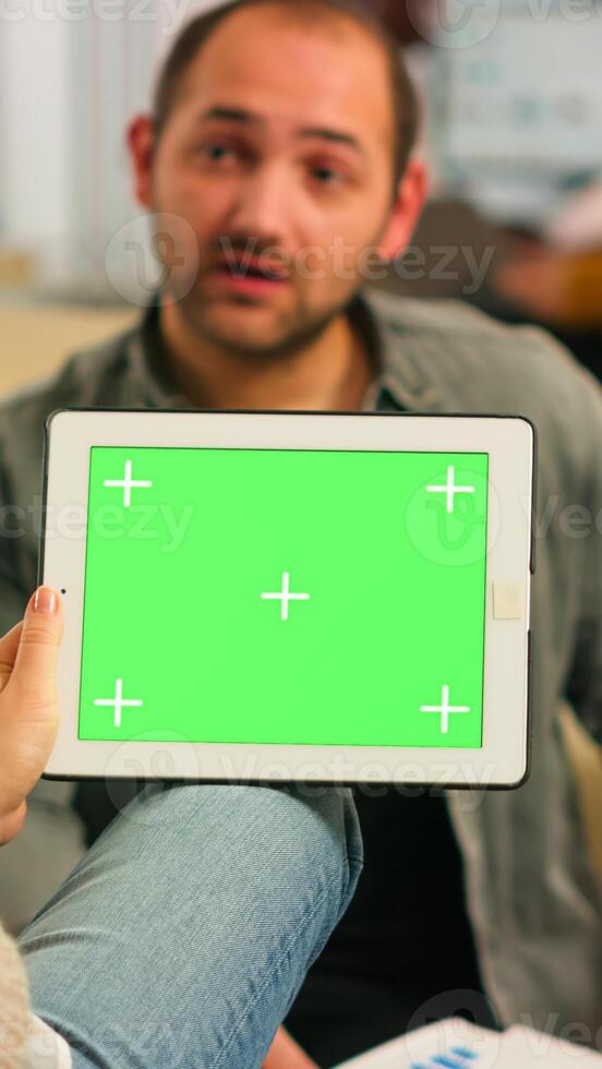 Over the sholder of manager woman sitting on couch talking with coworker holding tablet with green screen while diverse team working. Multiethnic people planning project on chroma key display photo