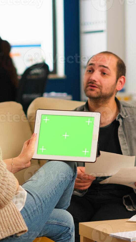 Back view of businesswoman sitting on couch using tablet with green screen talking with colleague while team working on background. Multiethnic coworkers planning project on chroma key display photo