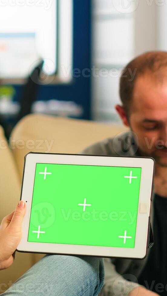 Business people sitting on couch analysing financial statistics, holding tablet with green screen while diverse team working on background. Multiethnic coworkers planning project on chroma key display photo