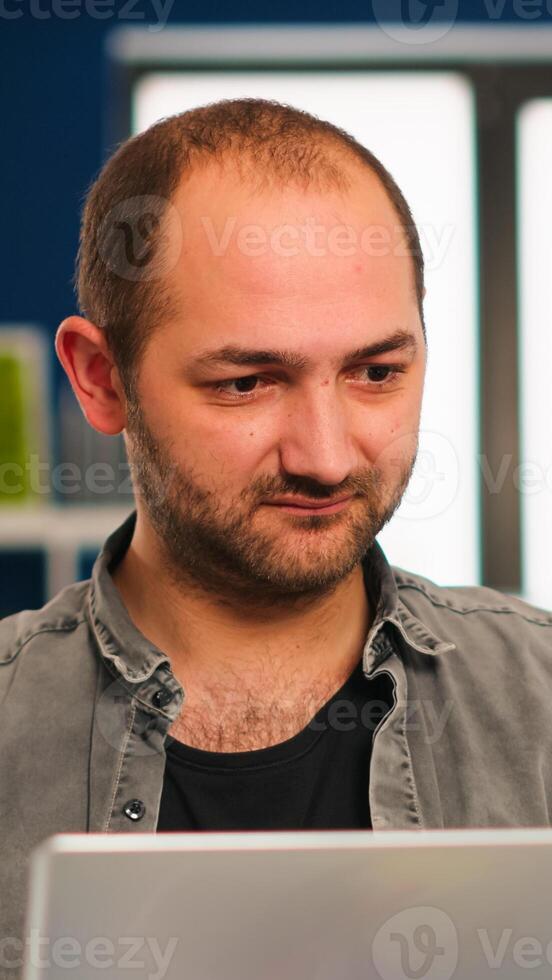 Enthusiastic business man writing a report sitting at table in modern startup business office sending mails typing on laptop, greeting colleague. Man working in financial company with diverse team. photo