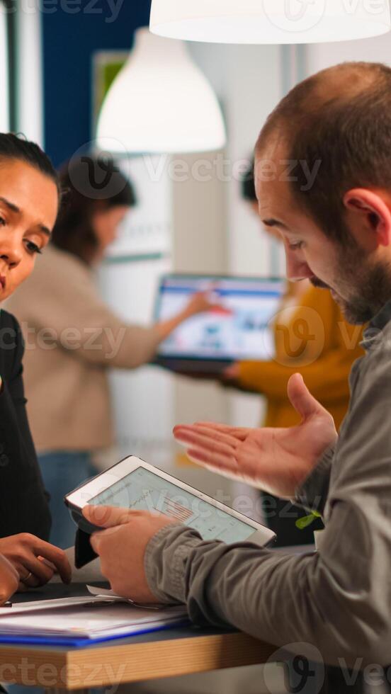 Close up of diverse business team checking graphs from tablet making new strategy. Manager explaining to multiethnic colleagues new financial project sitting at desk in moder startup office photo