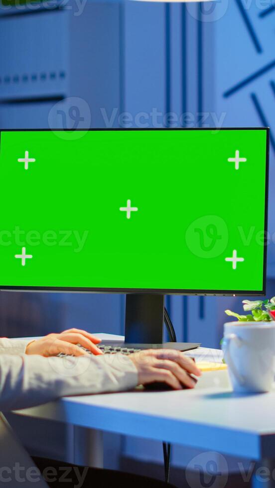Businesswoman looking at green screen monitor of computer sitting at desk in business office late at night. Freelancer watching desktop monitor display with green mockup, chroma key working overtime. photo