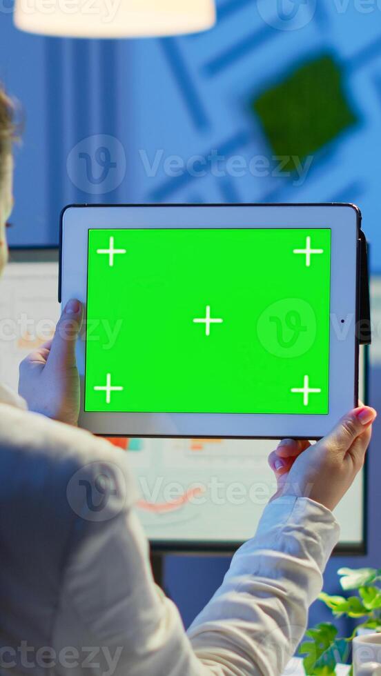 Businesswoman holding tablet with green screen monitor sitting at desk in business office late at night. Freelancer watching desktop monitor display with green mockup, chroma key working overtime. photo