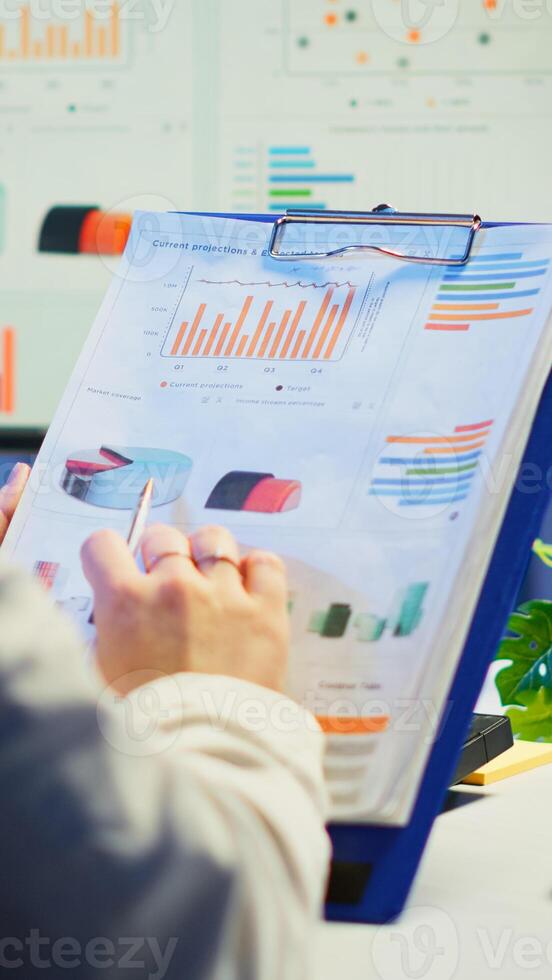 Close up of business woman holding clipboard with graphics and financial statistics working overtime in front of computer sitting in start-up business office. Busy employee using modern technology photo
