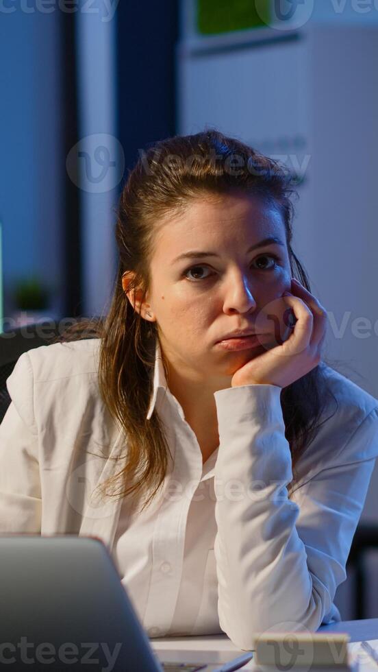 Exhausted businesswoman typing an email late at night and looking tired in camera sighing resting head in palm. Focused employee using technology network wireless doing overtime for job photo