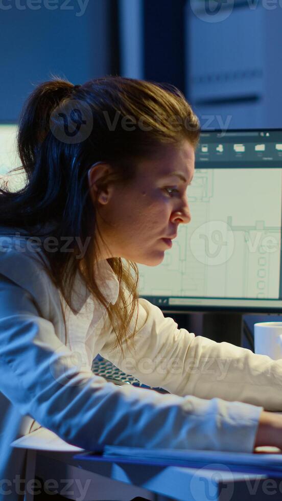 Focused woman architect checking and matching blueprints sitting at desk in front of computer late at night. Designer using arhitecture plans of buildings working overtime, creating and studying photo