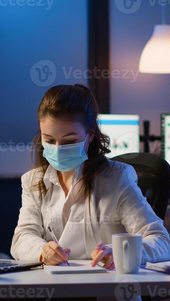 Manager woman with face mask working overtime in new normal business office checking team project, taking notes, analysing financial documents sitting at desk late at night during global pandemic photo