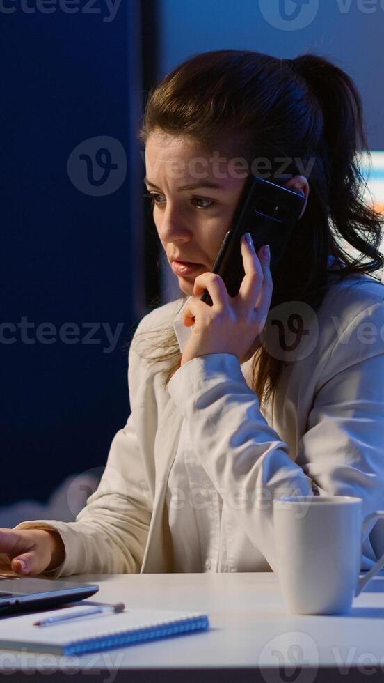 Woman employee speaking at phone while working at laptop late at night. Busy focused freelancer using modern technology network wireless doing overtime for job reading writing, searching taking break photo