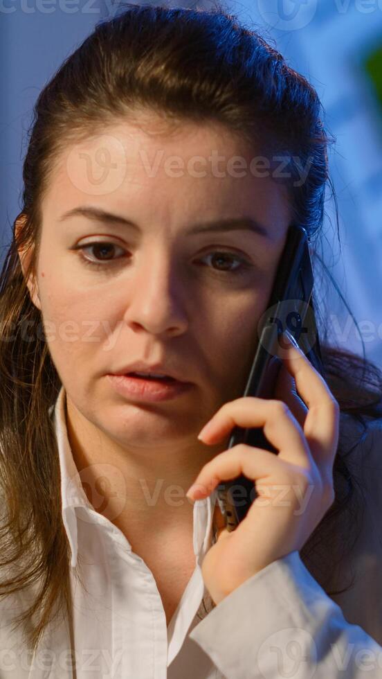 Concentrated businesswoman during phone call late at night working from business office discussing with coworker. Busy employee using modern technology network wireless doing overtime for job talking photo