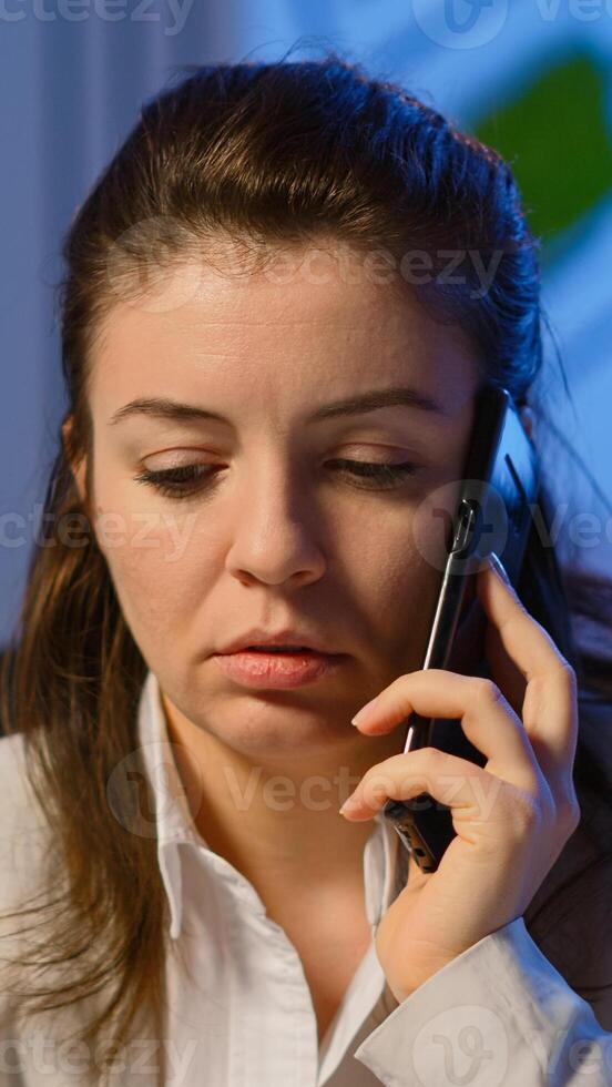 Tired freelancer woman having conversation on phone while working exhausted in business office late at night doing overtime. Focused employee using modern technology network wireless overworking photo