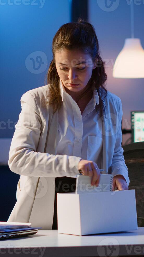 Sad fired businesswoman packing things in box being dismissed from job late at night. Unemployed woman closing laptop and leaving workplace office in midnight photo