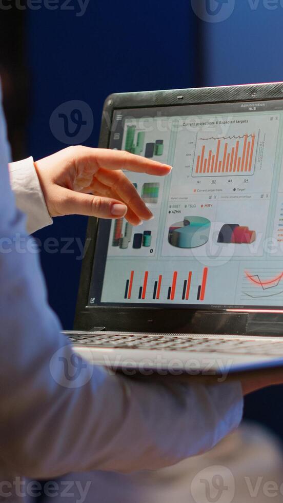 Back view of business woman analysing financial statistics holding laptop standing in start-up office late at night overworking. Busy focused employee using modern technology network wireless overtime photo