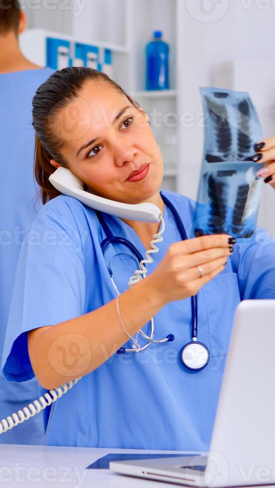 Physician nurse answering patient call by phone, looking at his radiographys, mri scan. Healthcare physician in medicine uniform, receptionist doctor assistant helping with phone concultation photo
