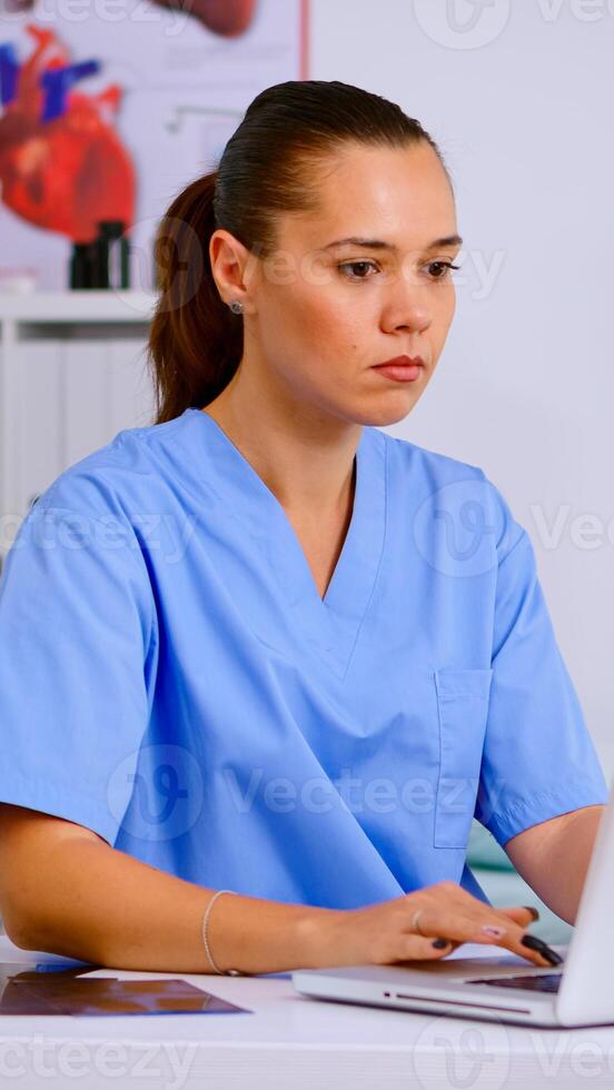 Medical practitioner typing patient health report on laptop in hospital office. Health care staff sitting at desk talking at phone in background, using computer in clinic looking at monitor, medicine. photo