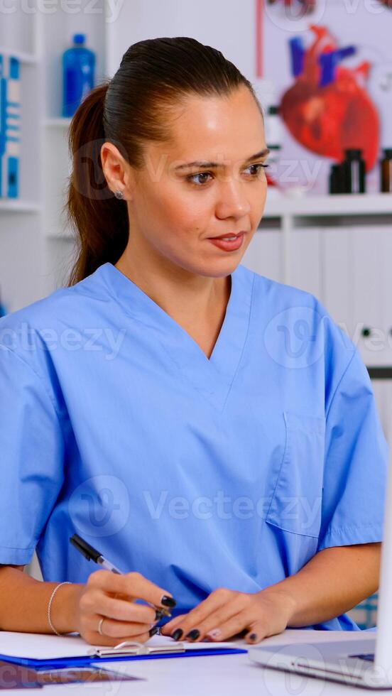 Doctor assistant in uniform working on laptop in modern hospital office. Therapist writing list of consulted patients online, making research, noting, taking useful information on clipboard photo