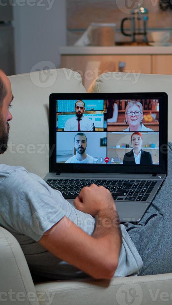Comfortable man relaxing on couch while having online business conference with collegues using laptop computer during quarantine. Focused adult talking on videocall use webcam late at night in kitchen photo