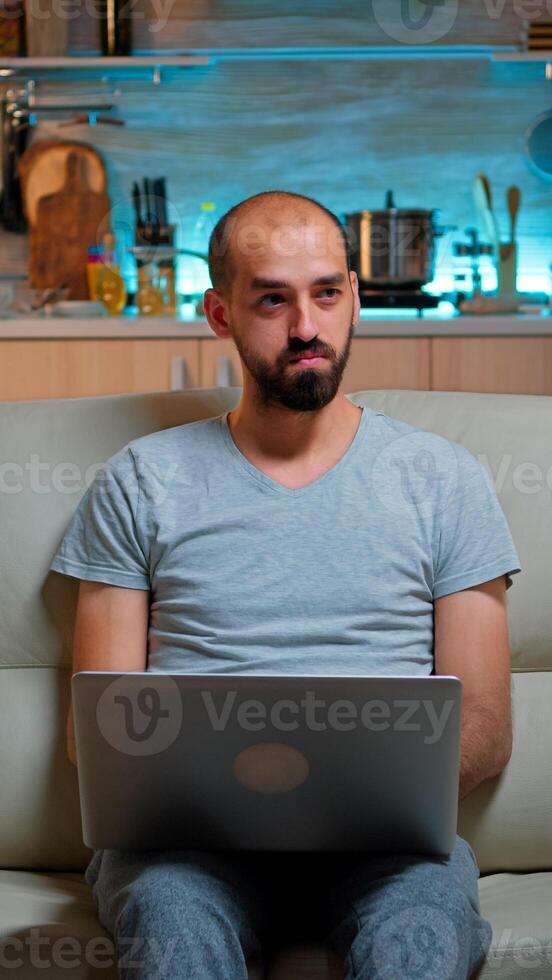Focused man sitting on sofa and writing online project on laptop late at night in living room. Caucasian male working from home while watching tv, browsing on internet using network technology photo