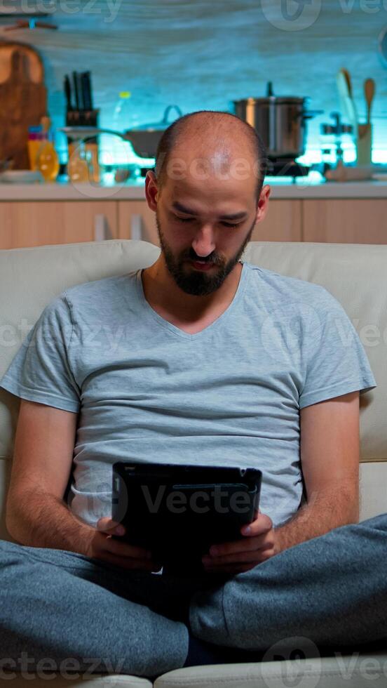 Tired man sitting alone on couch while browsing on internet using tablet computer with modern technology wireless. Caucasian male in pajamas relaxing in front of televion late at night in kitchen photo