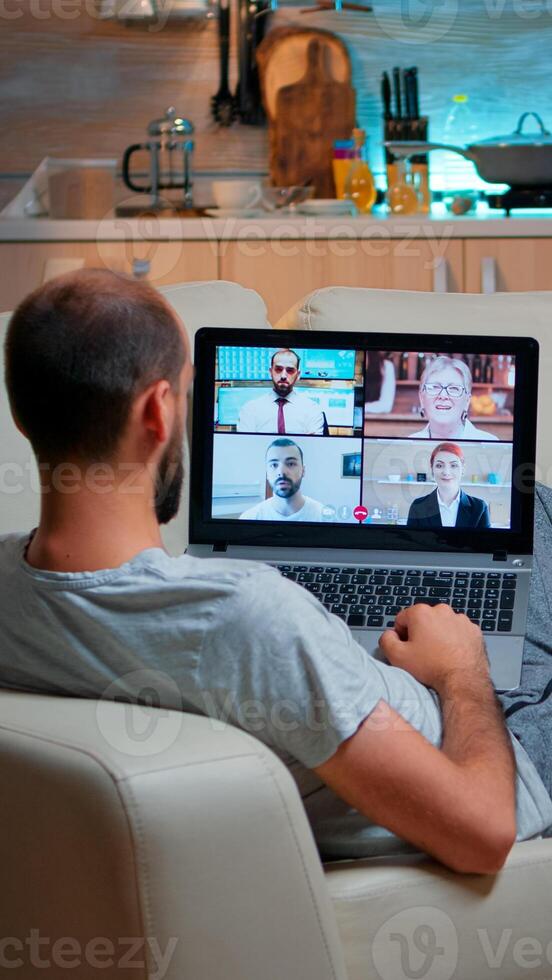 Caucasian male talking with collegues during call conference using laptop computer with modern technology wireless. Tired man relaxing on sofa late at night in kitchen photo