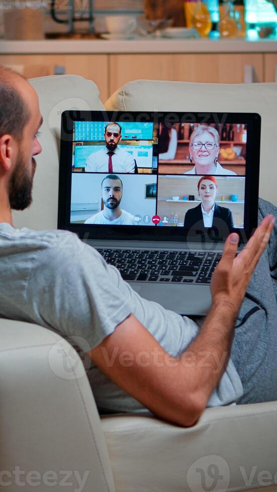 Tired man sitting comfortable on sofa while chatting with teammates about social communication during business videocall conference using laptop computer late at night in kitchen photo